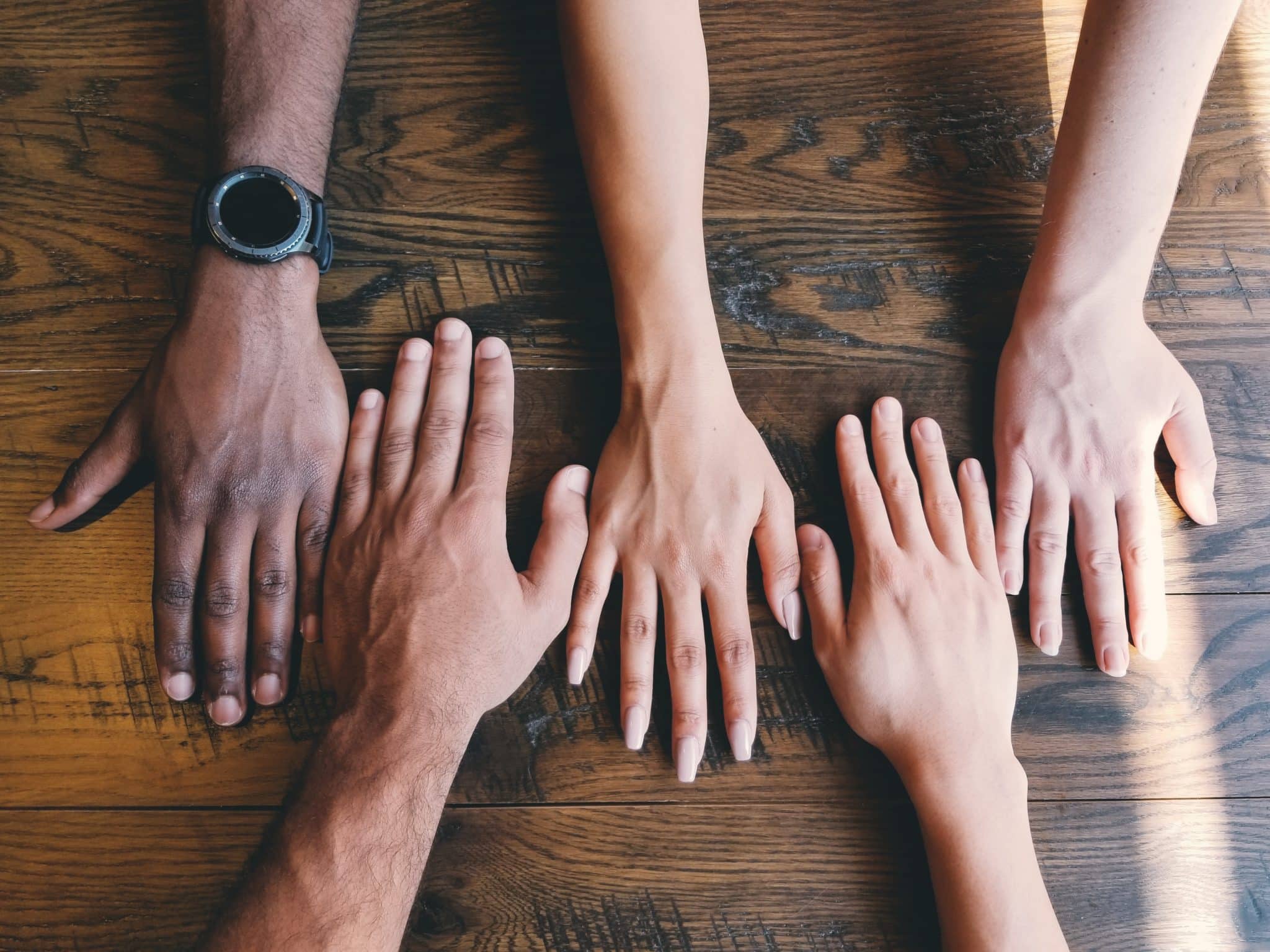 A top down photo of four different hands side by side on a table.  The hands are different ethnicities and genders all coming together to create a holistic solution.